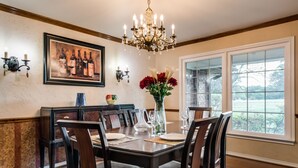 Une salle à manger avec une grande table dressée pour six personnes, un lustre, une œuvre d’art représentant une bouteille de vin encadrée et une fenêtre donnant sur un paysage verdoyant.