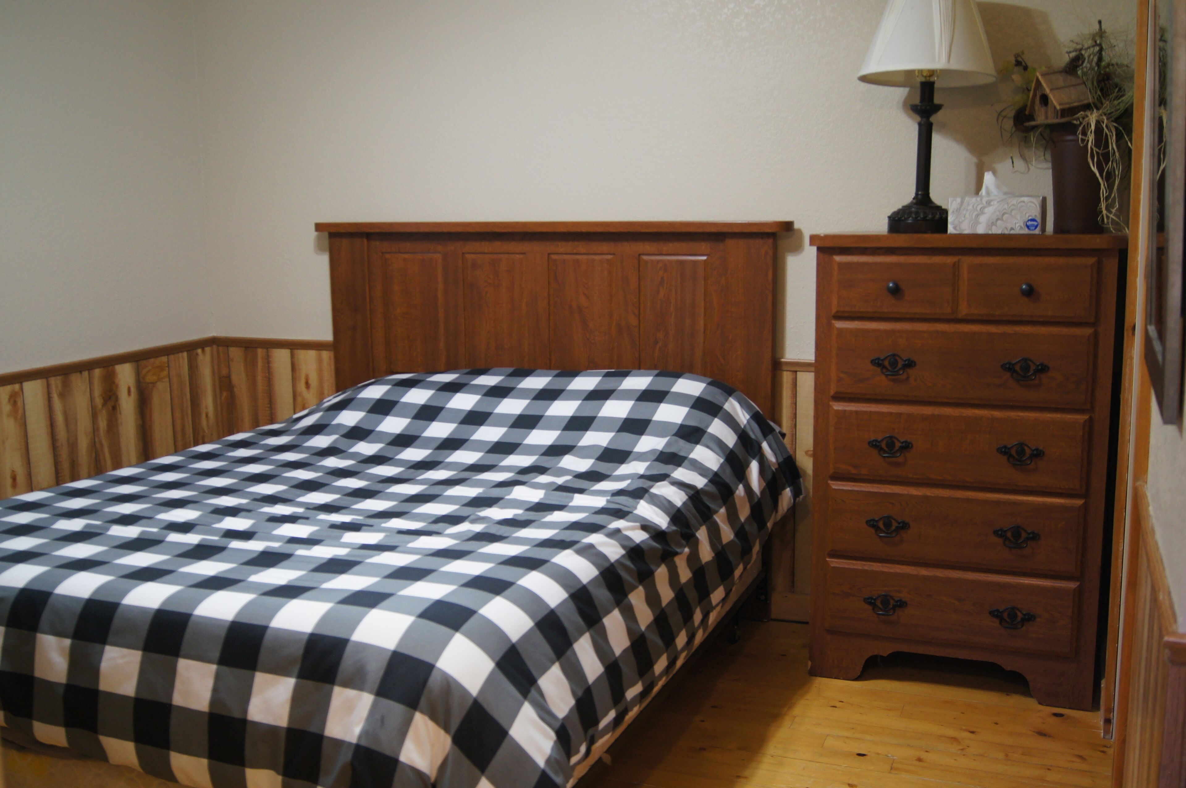 A bedroom with a checkered bedspread, a wooden headboard, a dresser with drawers, and a lamp on the dresser.