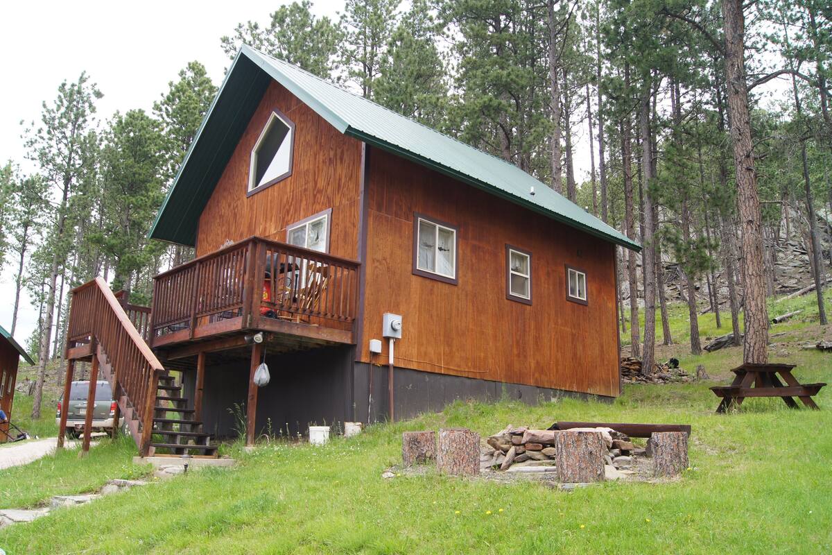 Una cabaña de madera con balcón, escaleras y una mesa de picnic en un área boscosa.