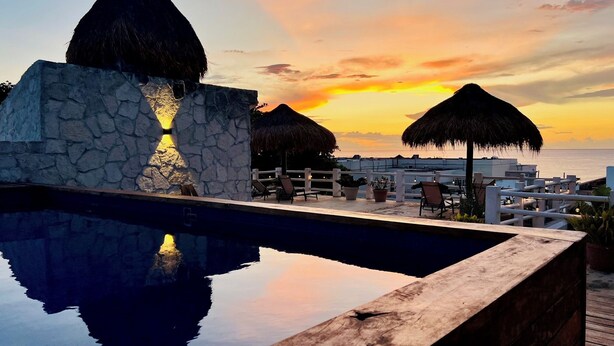 A pool with a stone wall and thatched umbrellas, overlooking a sunset over the sea. - Stunning Rooftop Villa with Breathtaking Caribbean Views & Large Private Terrace (San Miguel de Cozumel)