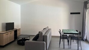 A modern living room with a sofa, a TV on a wooden cabinet, a dining table with chairs, and a window with curtains. - COZY GUEST HOUSE (BUEU)