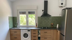 A compact kitchen with a washing machine, a sink, a window with blinds, a gas stove, and a built-in oven. - COZY GUEST HOUSE (BUEU)