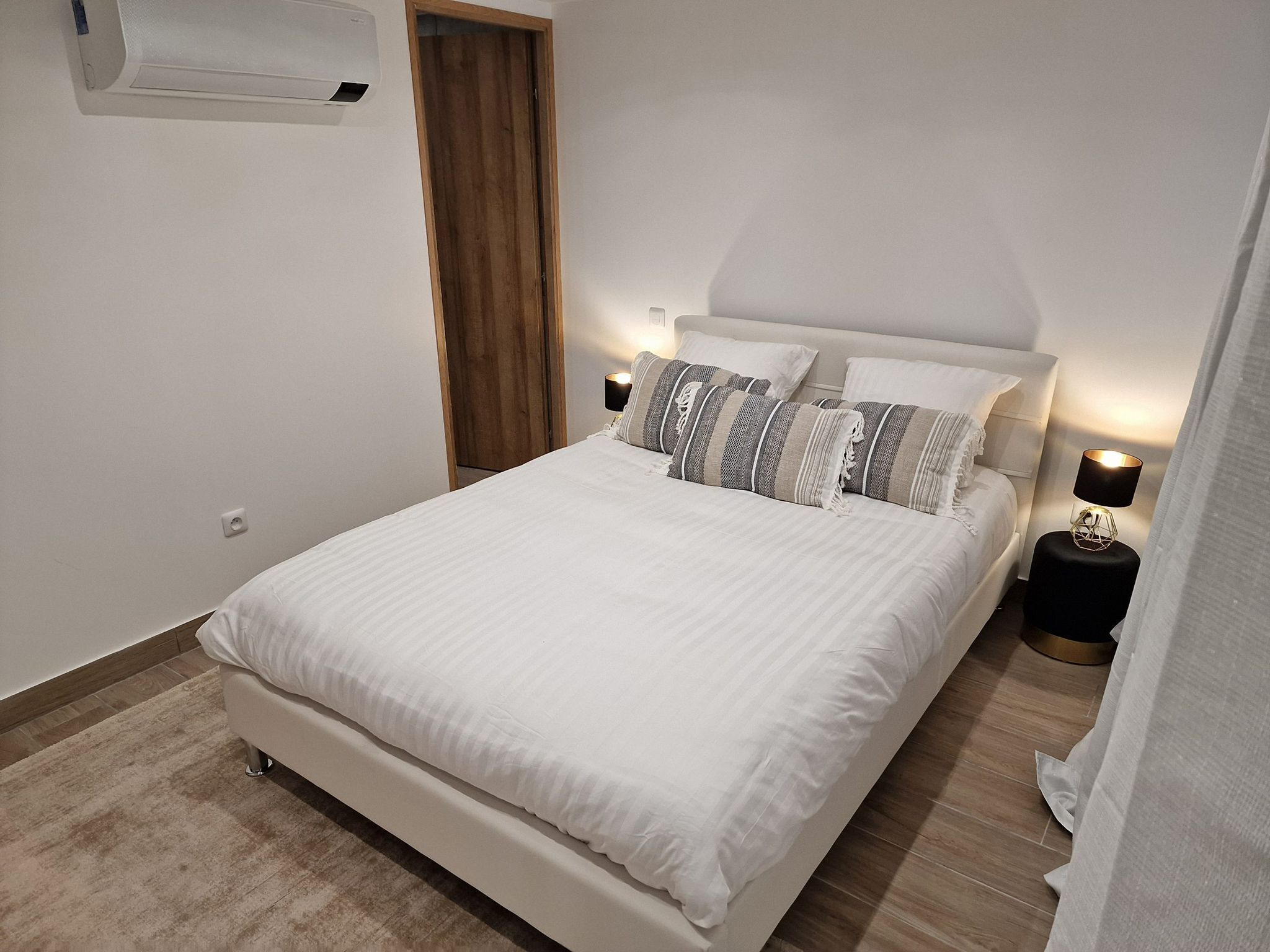 Una cama bien hecha con sábanas blancas y almohadas a rayas, una mesita de noche con lámpara, y una puerta de madera al fondo.