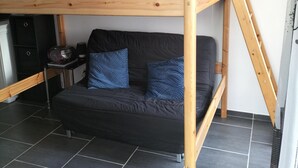 A bunk bed with a wooden frame, black mattress, and blue pillows. There is a small table with a radio and a cabinet below the bed. The room has dark tiled flooring and a white ceiling. - Studio near the beach (Criel-sur-Mer)