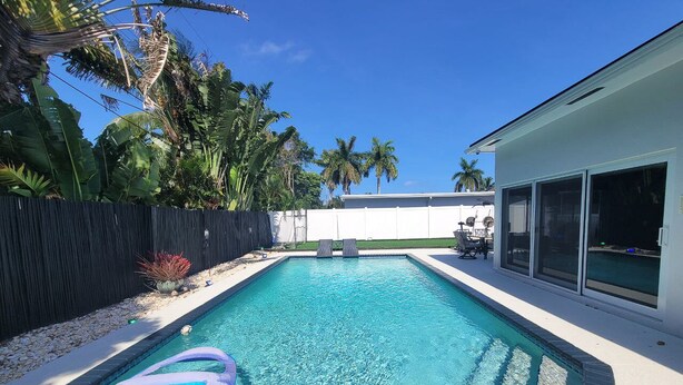 A rectangular swimming pool with clear blue water, surrounded by a black fence and palm trees.