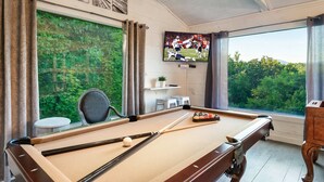 A pool table with a television displaying a sports game, a chair, and a view of greenery through large windows. - Hot tub cabin near Pigeon Forge, game room, mountain views (Sevierville)