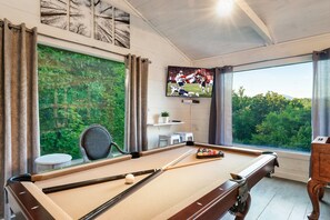 A pool table with a television displaying a sports game, a chair, and a view of greenery through large windows. - Summer Special! GAMES & PRIVATE HOT TUB! Pup-friendly! 4mi to Pigeon Forge (Sevierville)