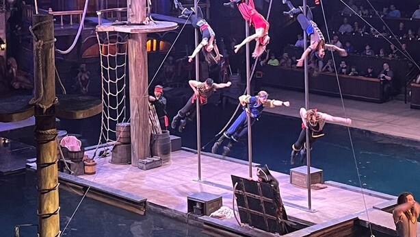 A circus performance with acrobats on poles and a pool in the background.