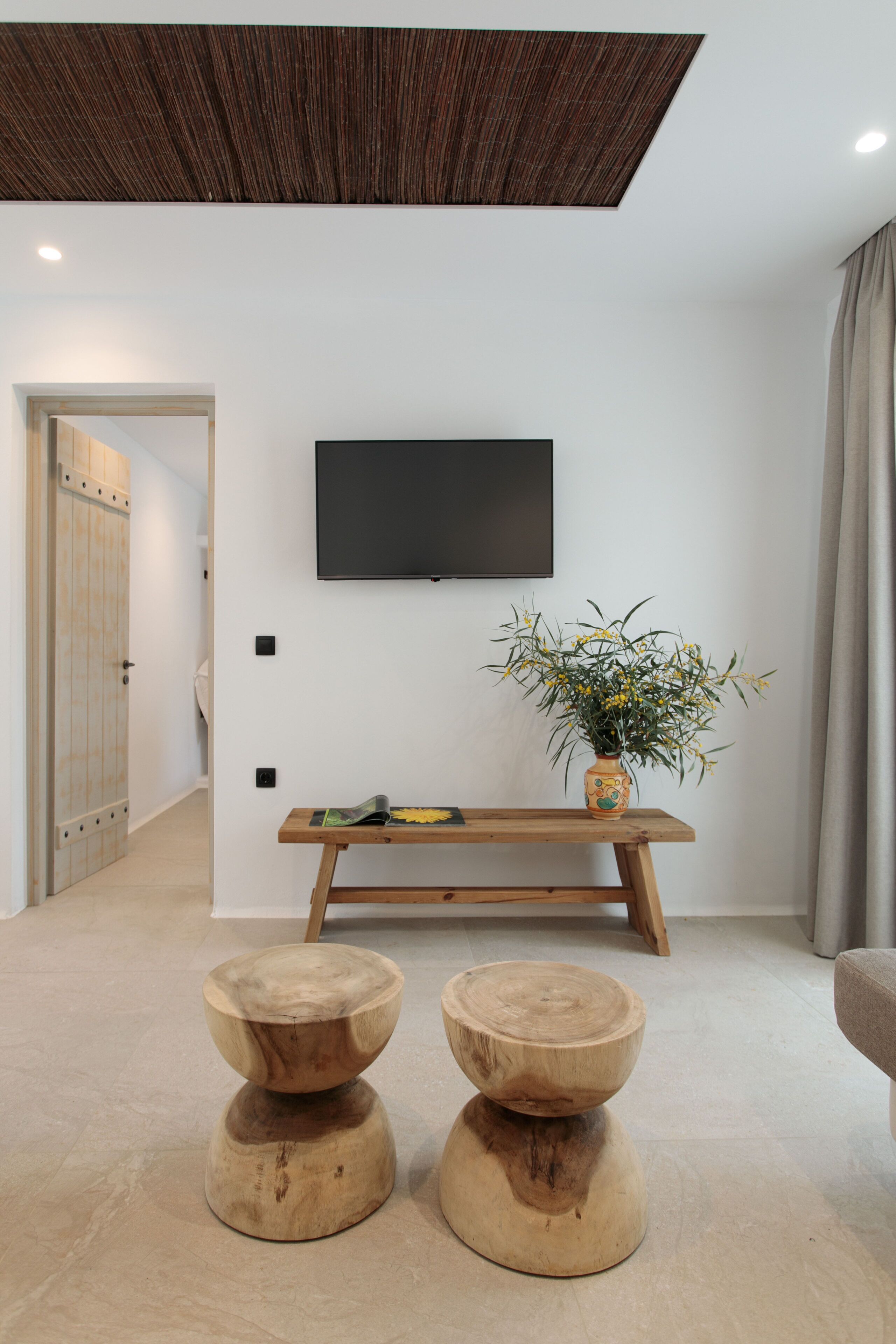 A modern living room with a wooden bench, a flat-screen TV mounted on the wall, and a wooden coffee table with a vase of flowers.