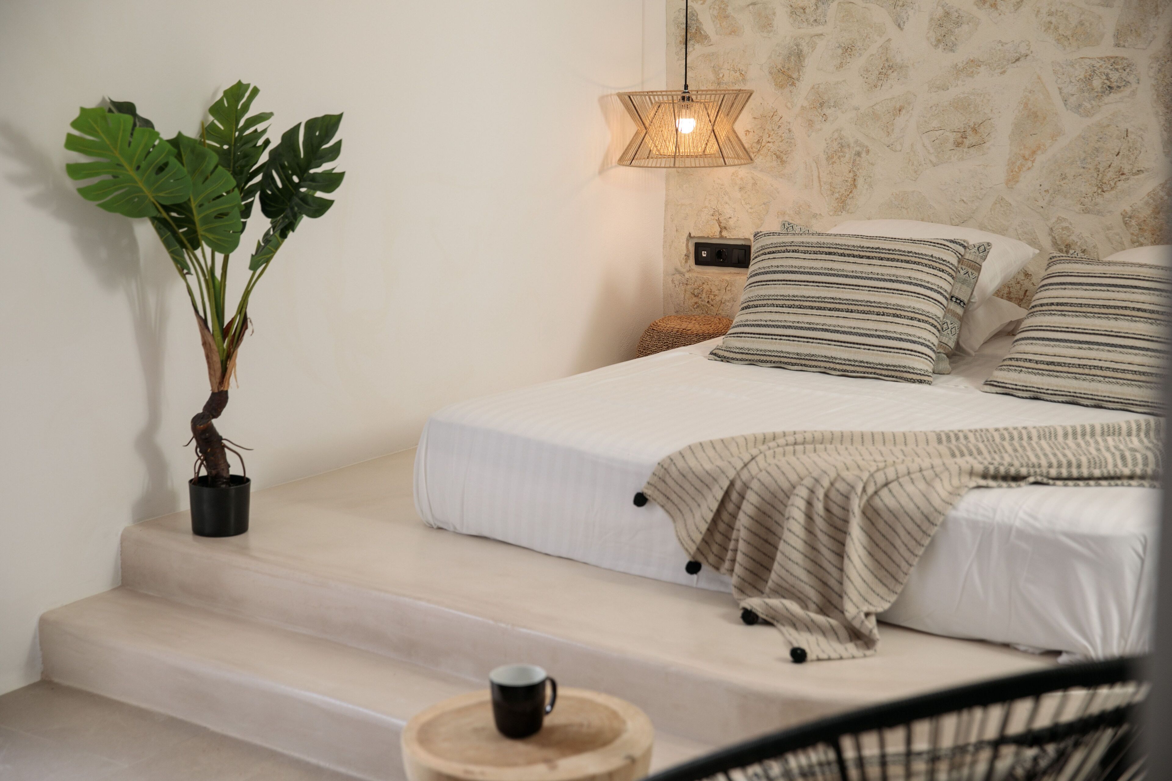 Un dormitorio con una cama, una planta en maceta, una lámpara de noche y un taburete de madera.