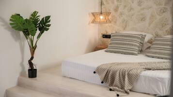 Un dormitorio con una cama, una planta en maceta, una lámpara de noche y un taburete de madera.