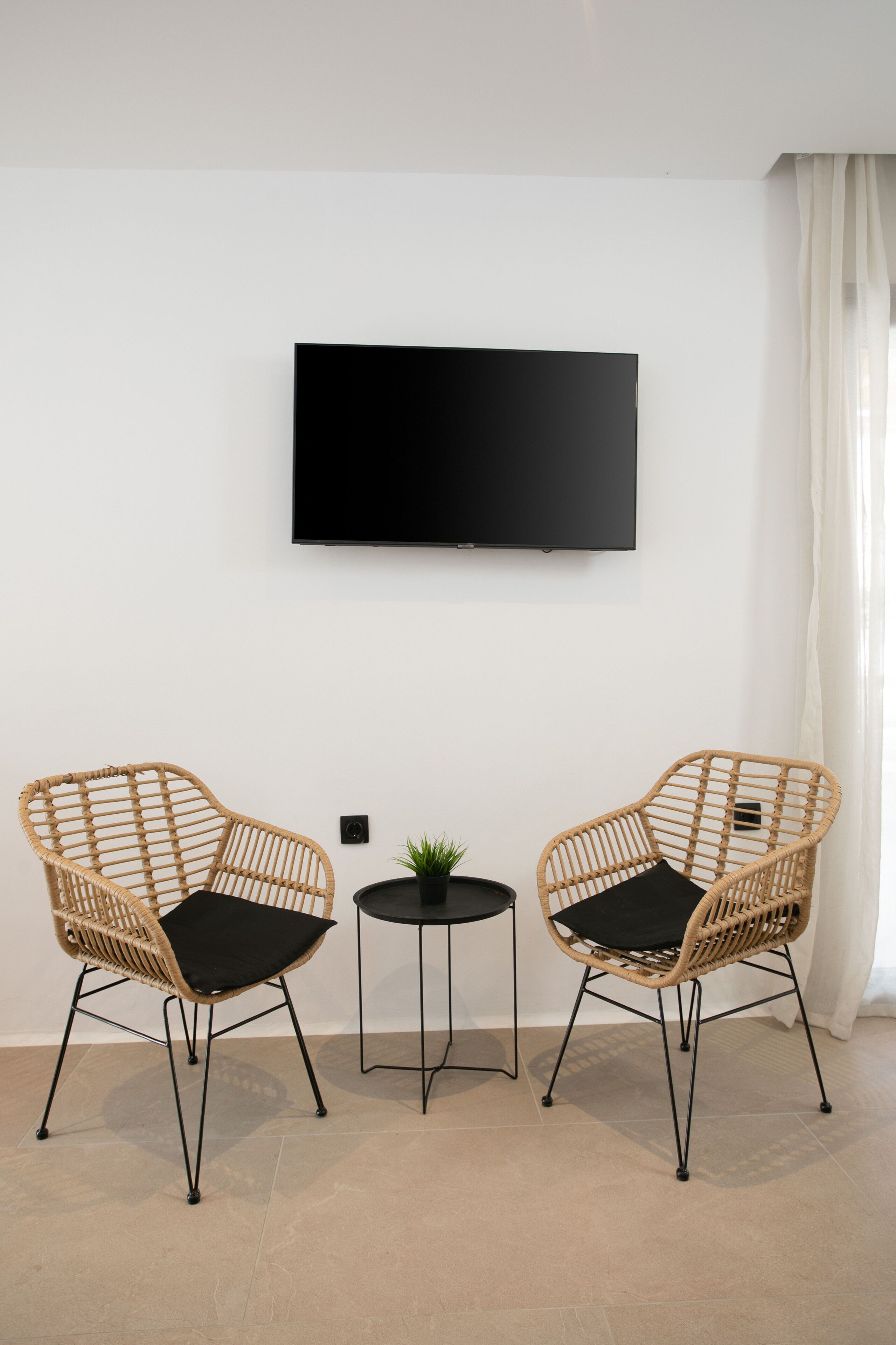Una sala de estar moderna con dos sillones de mimbre, una pequeña mesa redonda con una planta y un televisor montado en la pared.