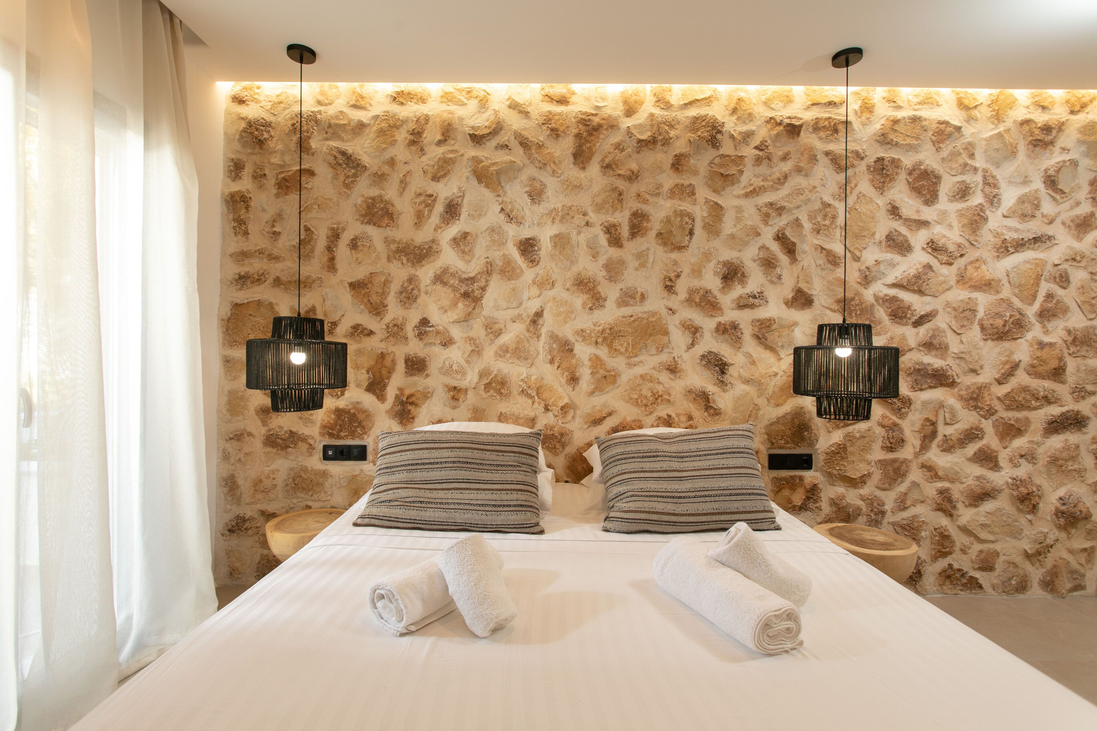 Una habitación de hotel moderna con pared de piedra, una cama bien tendida con almohadas a rayas y dos lámparas colgantes.