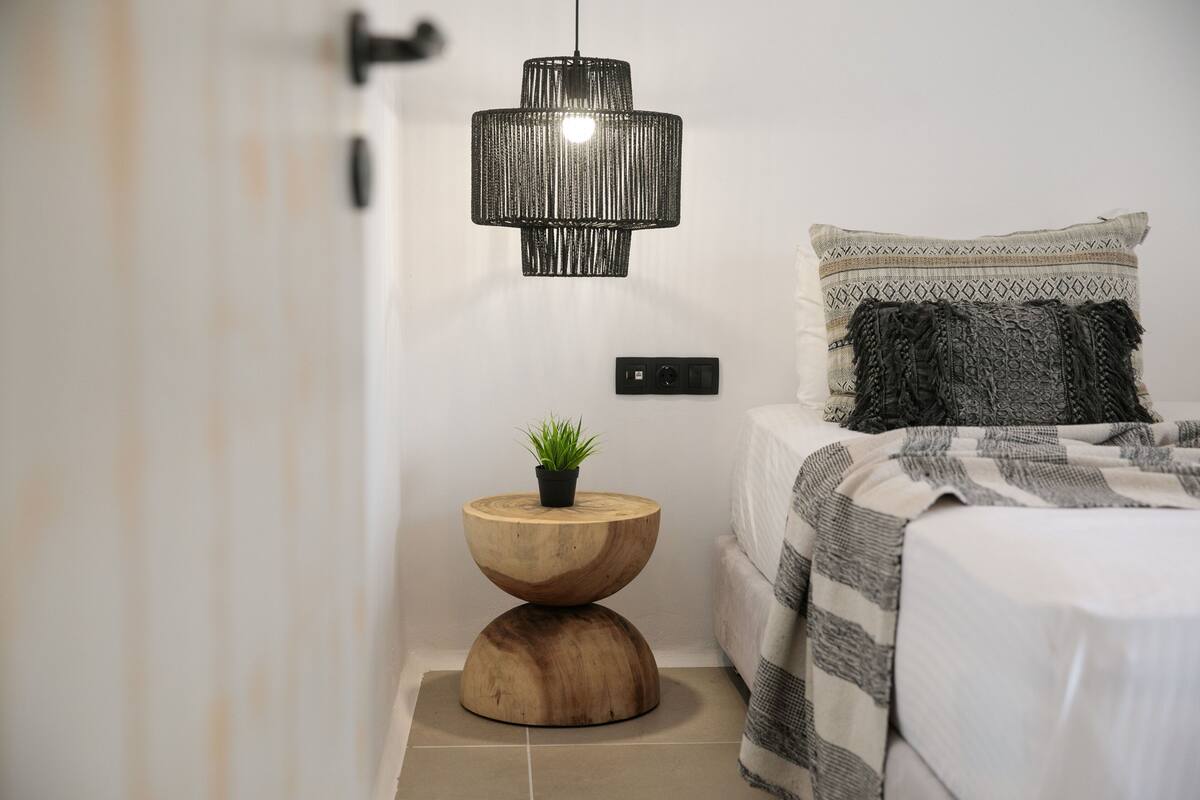 A bedroom with a bed, a bedside table with a plant, a hanging lamp, and a wall socket.