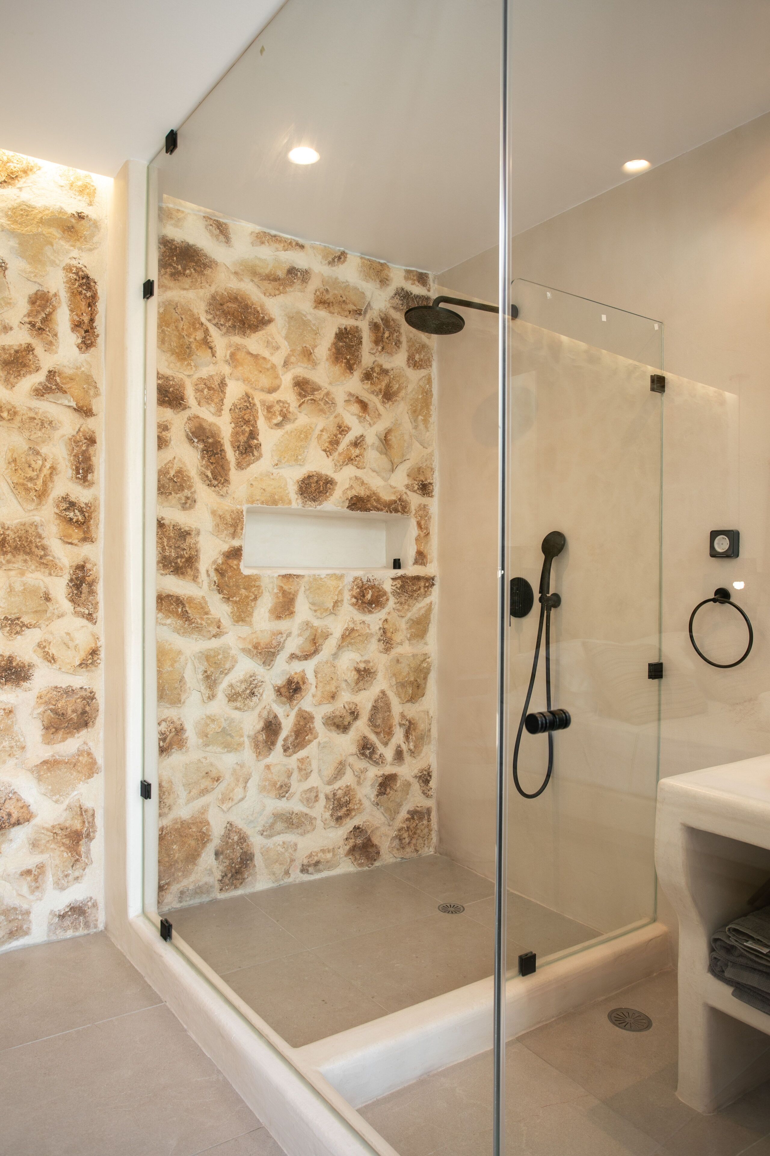 Un baño moderno con ducha envuelta en vidrio, pared de piedra y lavamanos blanco con toallero.
