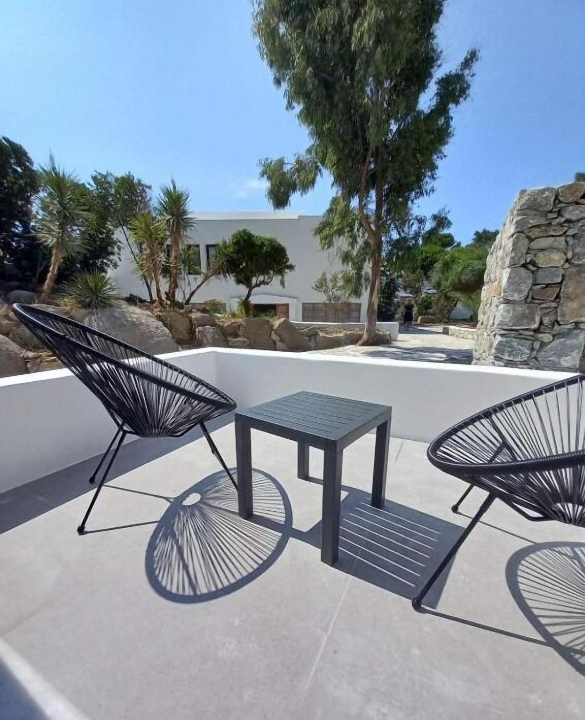 Un área de estar al aire libre con dos sillas de estructura metálica negra y una pequeña mesa cuadrada, rodeada por un muro blanco y un muro de piedra, con árboles y un edificio moderno al fondo.