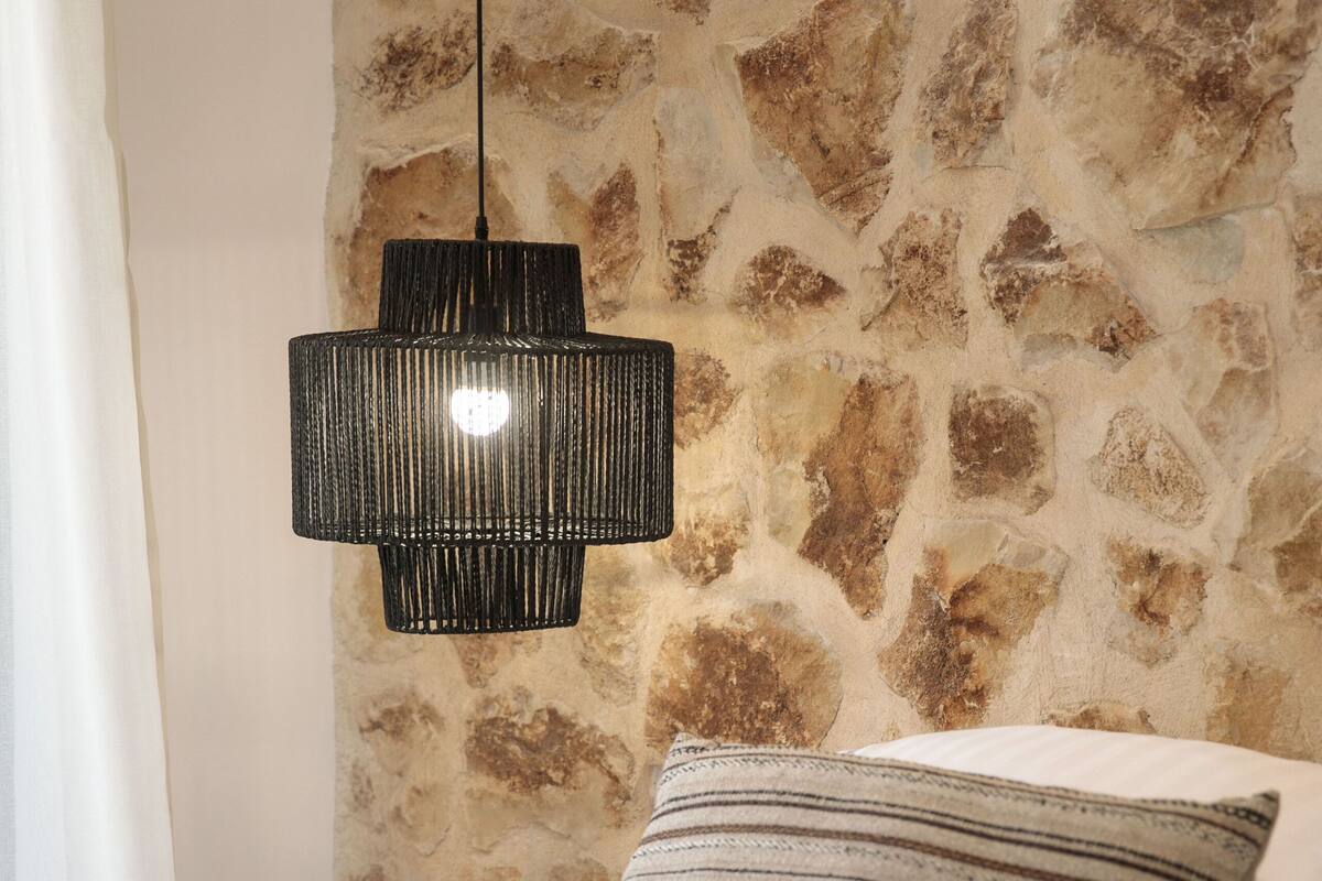 A bed with a striped pillow and a black pendant light against a stone wall.