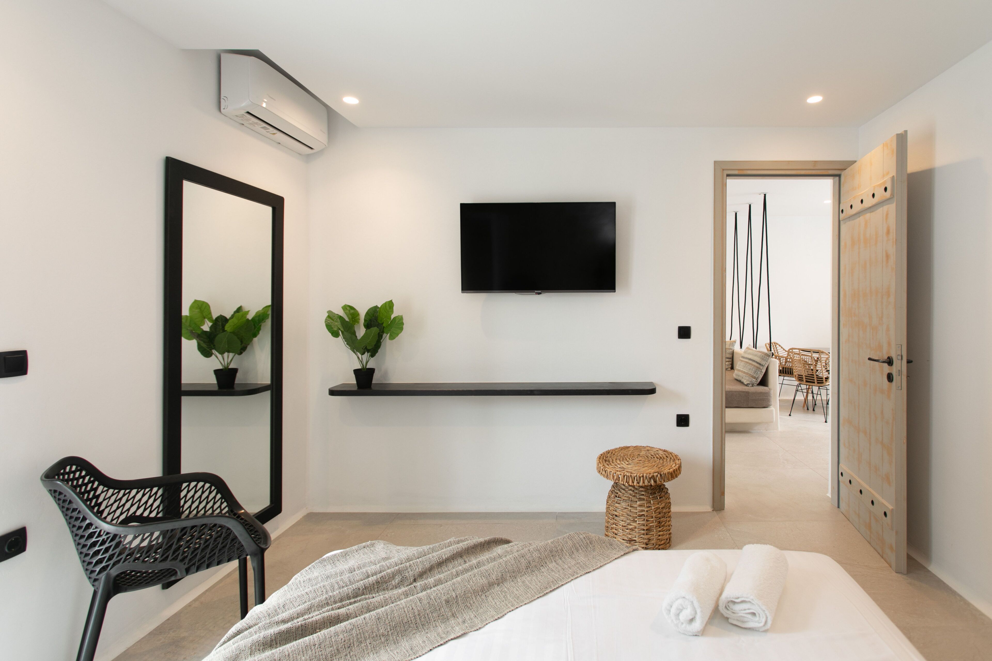 A modern hotel room with a bed, a TV mounted on the wall, a small plant on a shelf, and a wicker stool.