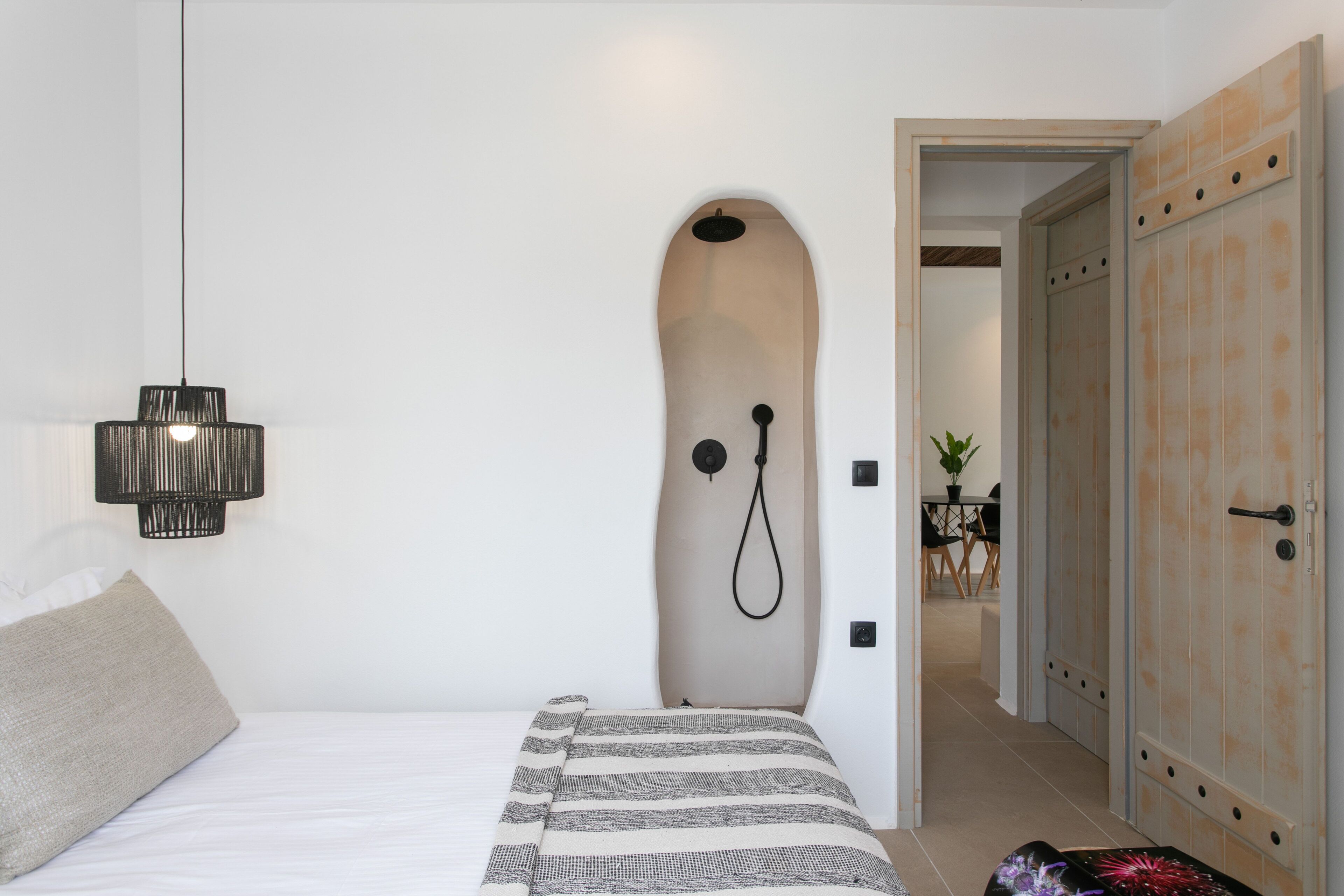 Une chambre avec un lit, une douche fixée au mur, un luminaire suspendu et une porte donnant sur une autre pièce.