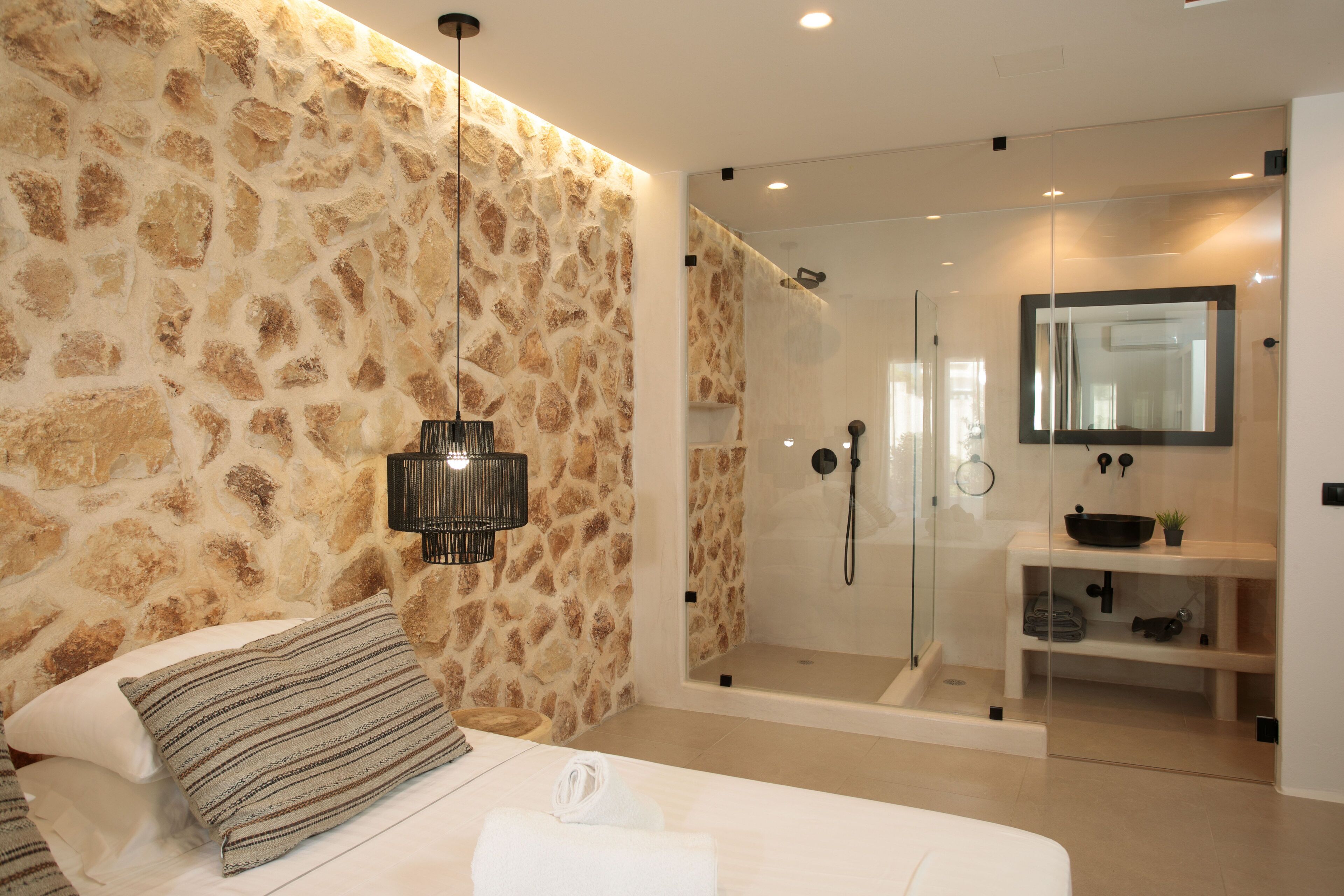 Une chambre moderne avec un mur en pierre, un lit agrémenté d’un coussin à rayures, une lampe de chevet et une douche séparée par des parois en verre.