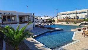 Une piscine avec des chaises longues, un palmier et un bâtiment sur lequel est apposée une enseigne indiquant : « Café et brunch ».