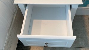 Una mesita de noche blanca con un cajón, un dispositivo electrónico negro encima y una pared azul con textura al fondo.