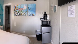 Un dormitorio con cama, mesita de noche, lámpara, reloj de pared, cuadro, microondas, refrigerador y basurero.