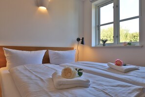 A hotel room with two beds, white linens, a wooden headboard, a wall-mounted lamp, and a window with a view of greenery.