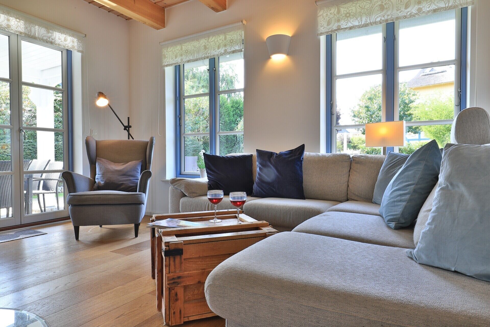 Un salon chaleureux avec un canapé, un fauteuil et une table basse. Il y a des fenêtres avec des rideaux, une lampe et une vue sur la verdure à l’extérieur.