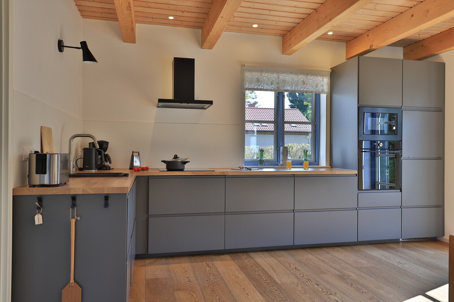 Une cuisine moderne avec des meubles gris, un plan de travail en bois, un four encastré et une fenêtre donnant sur une maison.