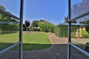 Uitzicht door een raam op een verzorgde tuin met een pad van stenen, een gazon en verschillende bomen en struiken.