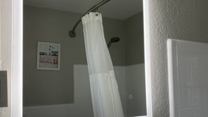 A bathroom with a shower, a white tiled wall, a framed picture, and a small shelf with toiletries. - Quaint Queen Deluxe (Daytona Beach)