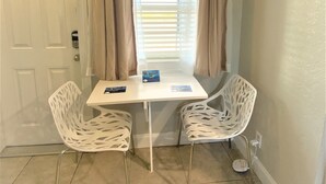 A small room with a white table and two white chairs, a window with blinds, and a door with a chain lock. - Quaint Queen Deluxe (Daytona Beach)
