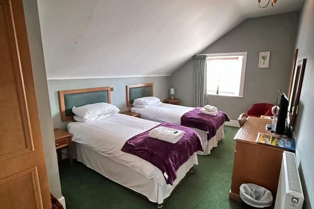 A hotel room with two beds, a desk, a chair, a radiator, and a window.