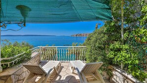 Eine Terrasse mit weißen Plastikstühlen und Steinboden, mit Blick auf das klare blaue Meer und eine ferne Küstenstadt.