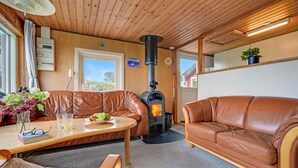 A cozy living room with a brown leather sofa, a wooden table with a plate of fruit, a black wood-burning stove, and a window with a view of greenery. - 1 bedroom awesome home in Allingåbro (Allingåbro)