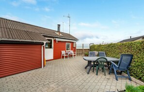 Un bâtiment rouge avec un toit en tôle ondulée, une porte blanche et une petite fenêtre. On y trouve une terrasse avec une table et des chaises, et un arbuste en arrière-plan.