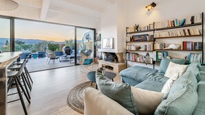 A modern living room with a large window, a sofa, a coffee table, and a bookshelf filled with books. - Villa Design D'architecte - 10 Personnes - Jacuzzi, Piscine ,tennis (Cassis)