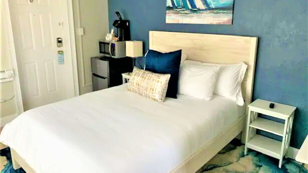 A neatly made bed with white linens and a blue pillow, a small bedside table with a lamp, and a painting of sailboats on the wall. - Zesty Queen Deluxe (Daytona Beach)