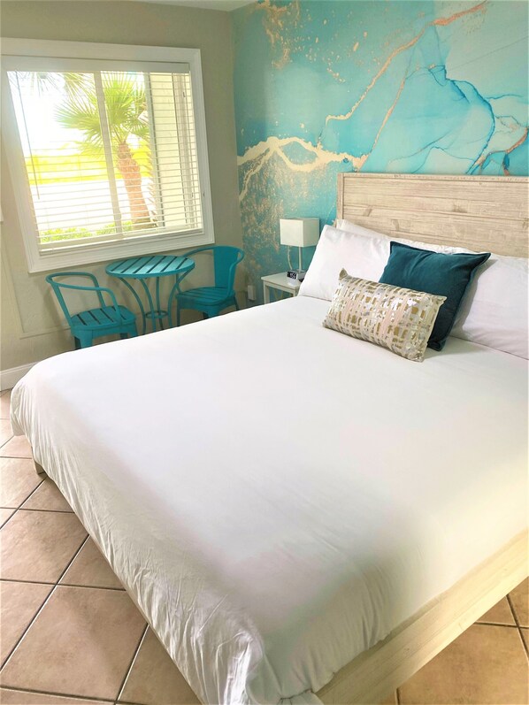 Un dormitorio con una cama grande, una mesita pequeña, dos sillas, una lámpara y un mural en la pared.