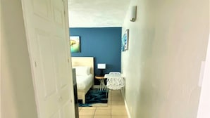 Un couloir avec une porte blanche, un lit avec du linge de lit blanc, une petite table avec une lampe et un tapis orné d’un motif ondulé.