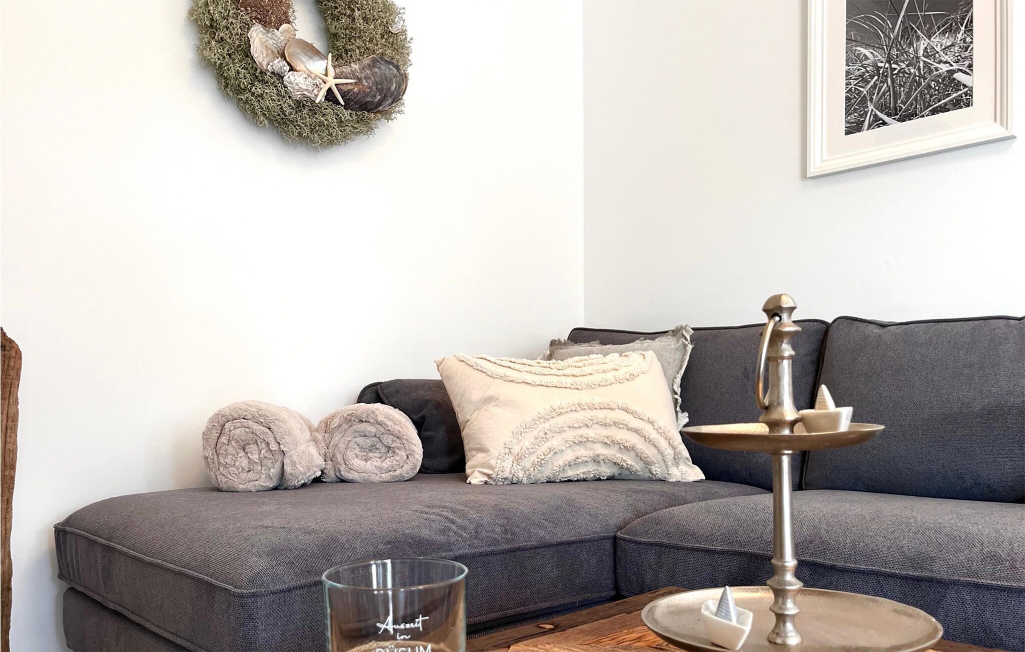 A living room with a grey sectional sofa, two rolled-up blankets, a metal table with a candle holder, a glass candle, and a framed picture on the wall.