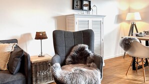 A cozy living room with a grey armchair, a small side table with a lamp, a wicker basket, and a white cabinet with shutters. - Awesome apartment in Büsum with sauna (Büsum)