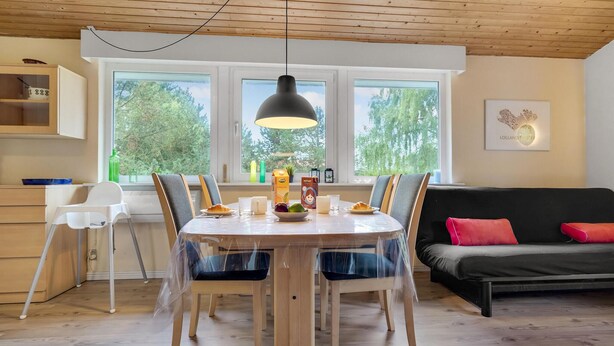 Een eethoek met een ronde tafel, stoelen en een zwarte hanglamp. Achtergrond is een zwarte bank met rode kussens.
