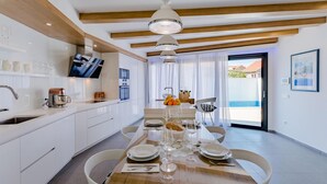 A modern kitchen with white cabinetry, a dining area with a wooden table set for four, and a view of a swimming pool through a glass door. - Lovely home in Sutivan (Sutivan)