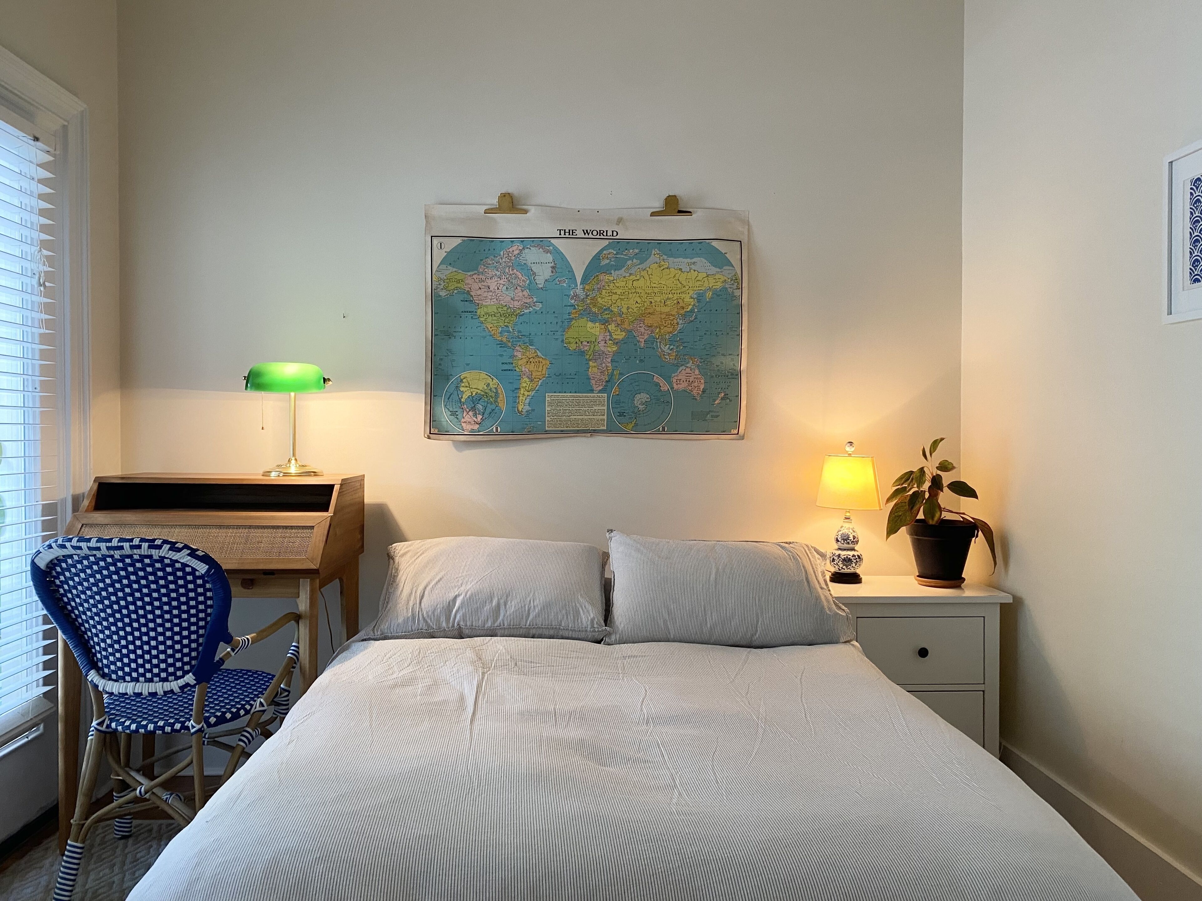 Ein Schlafzimmer mit einem Bett, einem Schreibtisch, einem Stuhl, einer Wandkarte, einer Lampe und einer Zimmerpflanze.