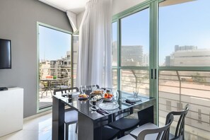 A modern dining area with a glass table set for four, large windows offering a city view, and a flat-screen TV mounted on the wall. - Mamad Elegant Sea-View Apt w Mamad by Sea N' Rent (Tel Aviv-Yafo)