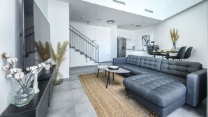 Un salon moderne avec un canapé d’angle gris, un téléviseur à écran plat, un coin repas avec une table et des chaises, et un escalier menant à un autre niveau.