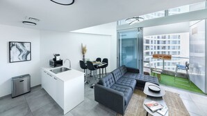 Un appartement moderne comprenant une cuisine, un coin repas et un salon. Il dispose d’une grande fenêtre offrant une vue sur un immeuble.