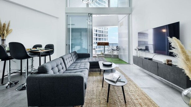 Un salon moderne avec un canapé d’angle gris, un coin repas avec des chaises noires, un téléviseur à écran plat et une grande fenêtre donnant sur un immeuble.